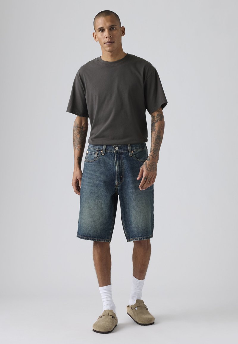 Homme tatoué portant un t-shirt gris foncé, un short en denim, des chaussettes blanches et des chaussures beige à enfiler, debout devant un fond clair uni.