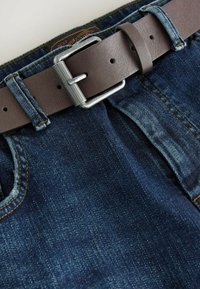 Jeans denim w ciemnoniebieskim kolorze, z brązowym skórzanym paskiem z metalową klamrą w kolorze srebra, przeszyciami oraz subtelnym wyblaskiem.