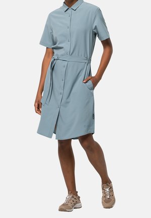 Osoba ubrana w jasnoniebieską sukienkę z krótkim rękawem zapinaną na guziki z paskiem, z jedną ręką w kieszeni, w połączeniu z beżowymi butami sportowymi.