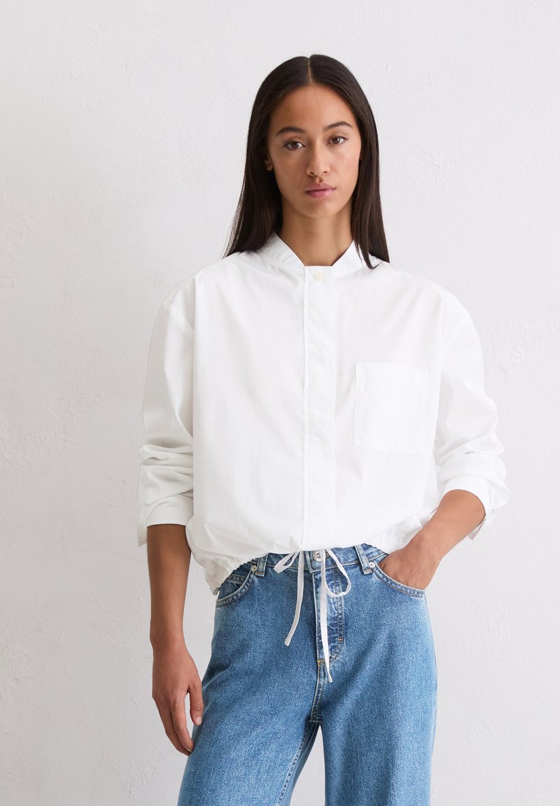 White button-up shirt with a stand collar, long sleeves, and a single chest pocket, paired with light blue, loose-fitting denim jeans.