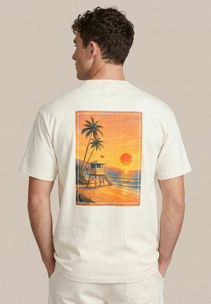 Man with curly dark hair, seen from back, wearing cream t-shirt featuring a sunset beach scene with palm trees and a lifeguard tower.