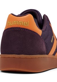 Närbild av ett lila och orange sneakershälen med en gul "hummel"-logotyp, texturerad sula och detaljer av mocka och nätfabric.