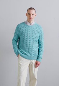 Pull en maille cable bleu clair avec des poignets et un ourlet côtelés, porté sur une chemise à rayures blanches et bleues, associé à un pantalon de couleur crème.