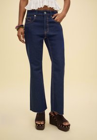 Ausgestellte blaue Jeans aus Denim mit orangefarbenen Nähten, fünf Taschen und einem hoch taillierten Design. Getragen mit braunen offenen Plattform-Highheels.