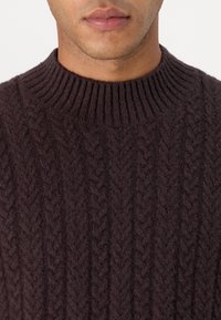 Pull tricoté marron foncé avec un col rond côtelé et un motif torsadé texturé sur tout le tissu. Matière douce en mélange de laine.