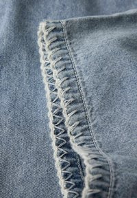 Svijetloplavi denim tkanina s krupnim prikazom cik-cak šava uz rub, s kontrastnom bijelom niti za naglasak.