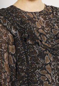 Blouse à motif sombre présentant un design imprimé serpent dans des tons de marron et de noir, avec un col froncé et un tissu texturé.