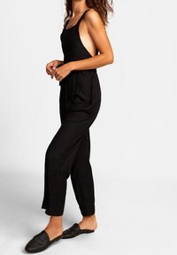 Mono negro con tirantes finos, de corte suelto y piernas anchas. Presenta bolsillos laterales y una tela texturizada. Combinado con sandalias negras.
