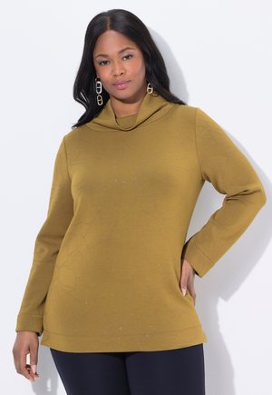 Femme portant un haut à manches longues jaune moutarde avec un motif floral subtil et un pantalon bleu marine, posant avec la main sur la hanche devant un fond blanc.