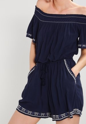 Kobieta w granatowym kombinezonie odsłaniającym ramiona, z białym haftowanym wykończeniem, elastyczną talią i kieszeniami, z rękami włożonymi do kieszeni.