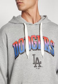 Sweatshirt gris avec capuche, présentant un graphique « DODGERS » en rouge et bleu dégradé, et le logo « LA » en noir. Cords de serrage blancs.