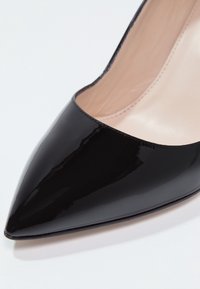 Detailní záběr na černou lakovanou botu na vysokém podpatku s špičatou špičkou a béžovým vnitřním podšívkou na bílém pozadí.