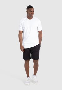 White cotton t-shirt with short sleeves, paired with black shorts. Model wears white sneakers, standing against a neutral gray background.