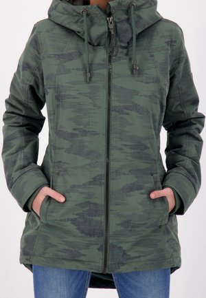 Gros plan d'une veste zippée camouflage verte avec capuche et cordons, portée avec les mains dans les poches avant et un jean bleu visible en dessous.