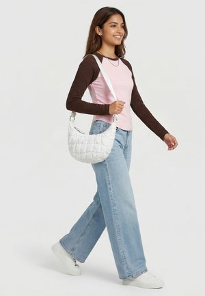 Junge Frau lächelnd, gehend in hellblauen Jeans mit weitem Bein, rosa-braunem langärmligen Hemd, weißen Turnschuhen, eine weiße gesteppte Umhängetasche tragend.