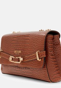 Borsa in pelle sintetica marrone con finitura a croco, dotata di una chiusura in tonalità oro, caratterizzata da una forma piatta, dettagli decorativi e la scritta "GUESS EST 1981" in rilievo sulla parte frontale.