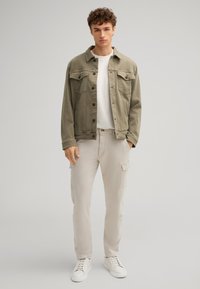 Giacca di jeans verde oliva con due tasche sul petto, abbinata a pantaloni cargo beige chiaro. Una t-shirt bianca a girocollo e sneakers bianche completano il look.