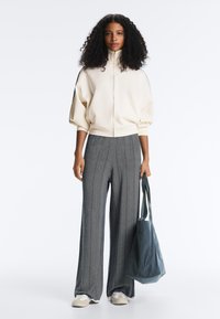 Frau mit lockigem Haar, die eine cremefarbene Jacke mit Reißverschluss, weite graue Hosen und weiße Turnschuhe trägt und eine große blaue Tragetasche hält.