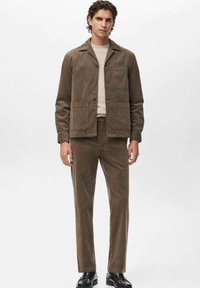 Giacca di corduroy marrone con colletto e tasche frontali, abbinata a pantaloni coordinati. Modello che indossa scarpe nere e una maglietta neutra sotto.