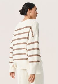 Pulover crem cu dungi orizontale maro, textură tricotată lejer, mâneci lungi și un design cu umăr lăsat. Tiv ribbed accentuat.