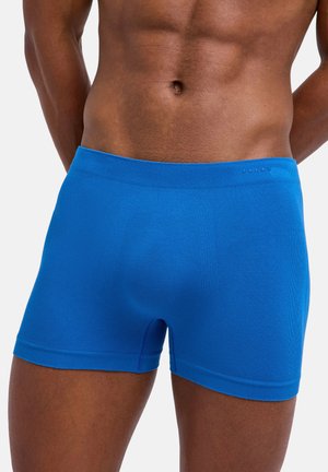 Atletyczny męski tors w jasnoniebieskich, bezszwowych bokserkach, stojący na jednolitym jasnym tle, z rękami za plecami.