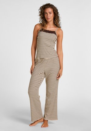 Femme aux cheveux bouclés debout pieds nus portant un haut sans manches beige et marron rayé et un pantalon large assorti.