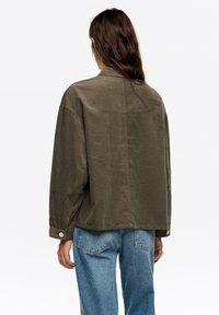 Zielona oliwkowa, teksturowana kurtka o lekko luźnym kroju, zapinana na guziki przy mankietach, z opuszczonymi ramionami, zestawiona z jasnoniebieskimi dżinsami.
