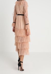 Kvinna iklädd en persikofärgad spetsklänning med lager i midi-längd, svart kant och svarta högklackade ankelbandsskor, stående med ryggen vänd mot kameran.