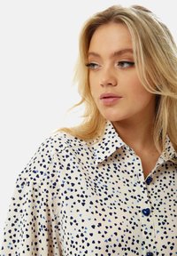 Button-up shirt made of lightweight fabric, featuring a cream background with clustered blue heart patterns and blue buttons.
