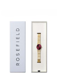 Montre dorée avec un cadran rond rouge, bracelet en or à stries, placée dans une boîte gris clair étiquetée "ROSEFIELD." Design minimaliste.