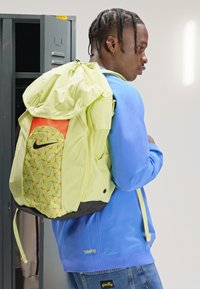 Rucsac galben neon cu un accent portocaliu; are un model geometric pe o parte, bază neagră și bretele ajustabile pentru umeri.