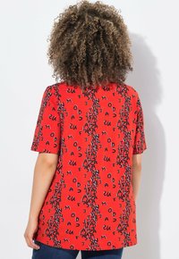 Camiseta roja de manga corta con un patrón de texto negro repetido. Corte recto y ajuste relajado. Tejido ligero y suave.