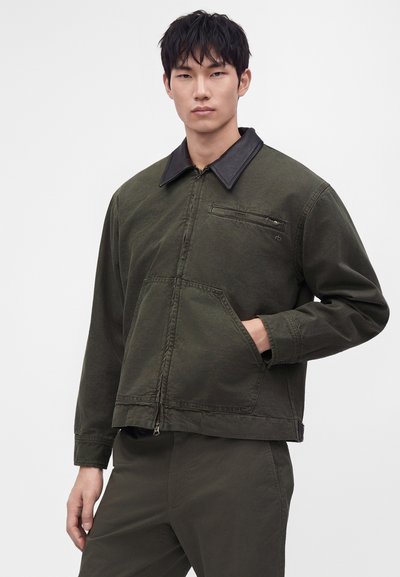 Jeune homme portant une veste vert olive avec col noir, fermeture éclair devant, poche poitrine et main dans la poche latérale, debout sur un fond uni.
