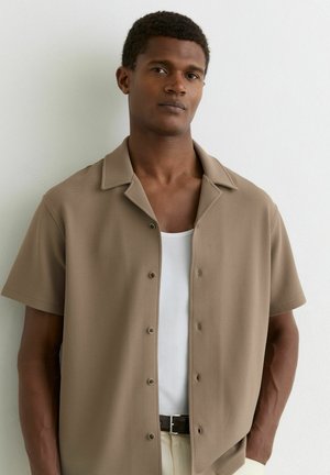 Lühikeste lokkis juustega mees kannab beeži lühikeste varrukatega särke valge altseelikuga, seisab valge seina taustal.