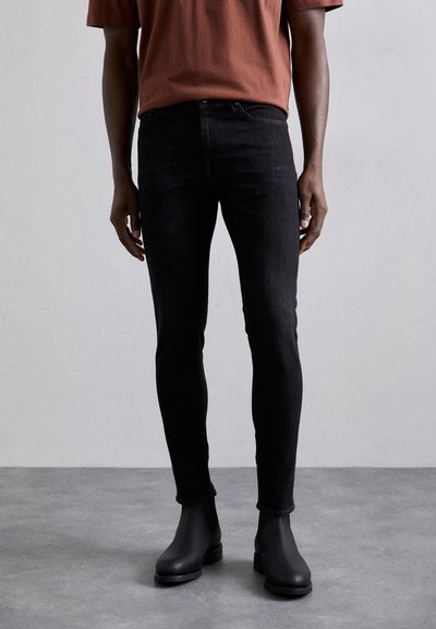 Jeans negri, mulati, din denim, cu un design clasic cu cinci buzunare, asortați cu ghete negre până la gleznă și un tricou de culoare roșie-ruginie.