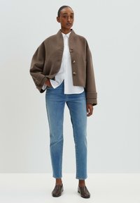 Veste en laine brune courte avec un col montant et des boutons ronds, portée sur une chemise blanche, associée à un jean bleu clair et des chaussures brunes.