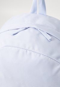 Világoskék szövet hátizsák, texturált felülettel, ívelt tetejű, zsinóros zárással és hurok alakú fogantyúval a tetején.