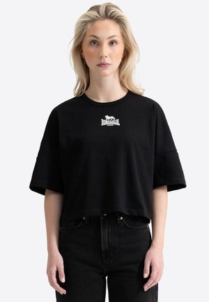 Blond žena oblečená v čiernom oversized krátkom tričku Lonsdale London a čiernych rifle s vysokým pásom stojaca na bielom pozadí.