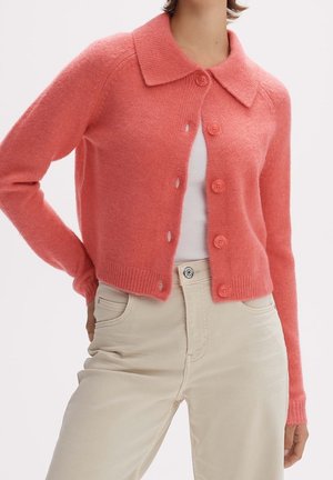 Femme portant un cardigan boutoné rose corail sur un haut blanc, assorti à un pantalon beige taille haute, sur un fond uni.