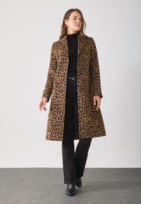 Luipaardprint jas met een kraag, knoopsluiting en lange mouwen, gecombineerd met zwarte broek en enkellaarsjes tegen een neutrale achtergrond.