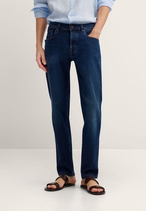 Mann trägt dunkelblaue Jeans, hellblaues hochgekrempeltes Hemd und braune Ledersandalen, steht mit einer Hand in der Tasche.