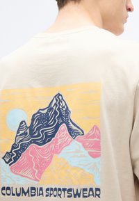 Μπεζ βαμβακερό t-shirt με πολύχρωφο γραφικό βουνό σε μπλε, ροζ και κίτρινο, με το κείμενο «COLUMBIA SPORTSWEAR» στο κάτω μέρος.