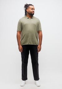 Άνδρας με dreadlocks που φοράει πόλο ελιάς, μαύρο παντελόνι και λευκά αθλητικά παπούτσια, στέκεται μπροστά σε απλό λευκό φόντο, κοιτάζοντας δεξιά.