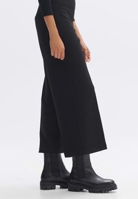 Pantalon noir à jambes larges en tissu texturé, associé à des bottines noires brillantes à semelle épaisse et fermetures éclair latérales.
