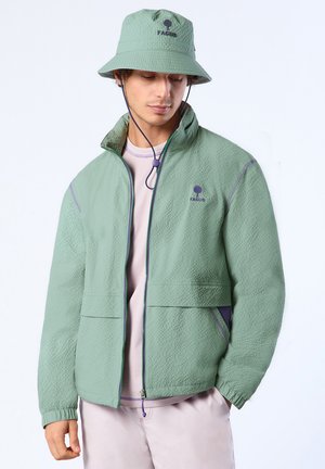 Jeune homme portant une veste zippée texturée verte et un chapeau bob assorti avec une sangle sous le menton, regardant vers le bas, les mains dans les poches.