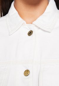 Giacca in denim bianco con colletto piegato, caratterizzata da due bottoni dorati e dettagli di cuciture a contrasto sul davanti.