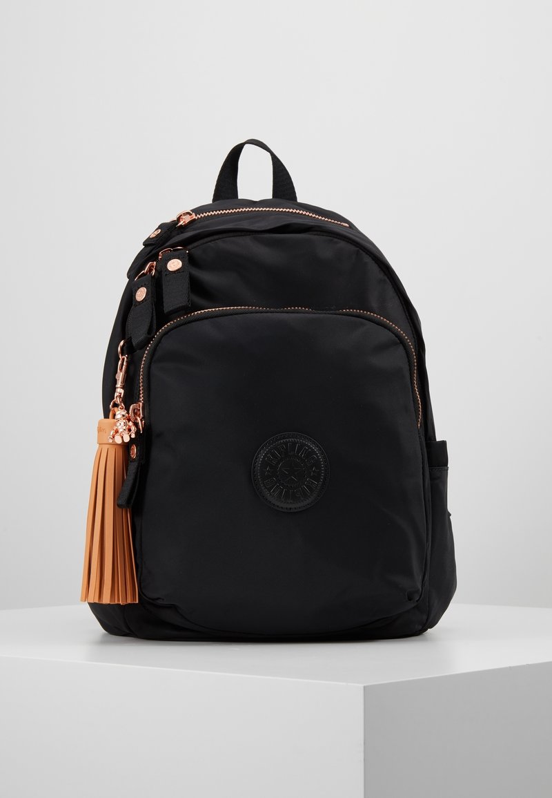 Sac à dos compact noir avec une grande poche avant, des fermetures éclair en or rose, un porte-clés à pompon beige et un écusson rond avec logo sur le devant.