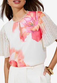 Floral blouse with white fabric, bright pink flowers, and sheer polka-dot sleeves. Features a round neckline with a delicate trim.
