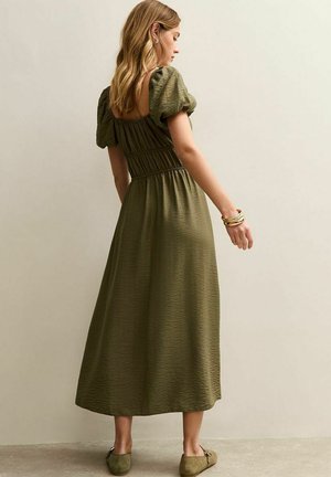 Vrouw staand opzij gekleed in een olijfgroene midi-jurk met pofmouwen en platte schoenen, tegen een effen beige muur.