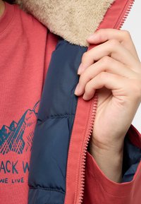 Veste rouge avec une douce doublure en fausse fourrure beige, un intérieur bleu et une fermeture éclair rose visible. Portée par-dessus une chemise corail avec un motif de montagne.